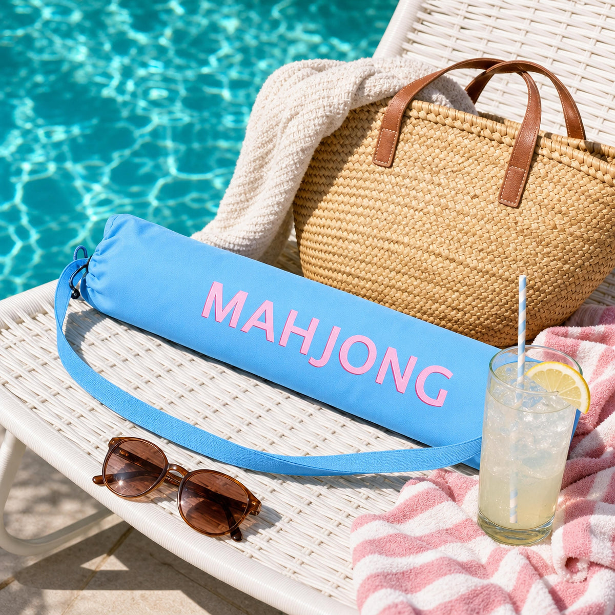 Blue mahjong mat carry bag on a poolside lounge chair with a straw tote and lemonade